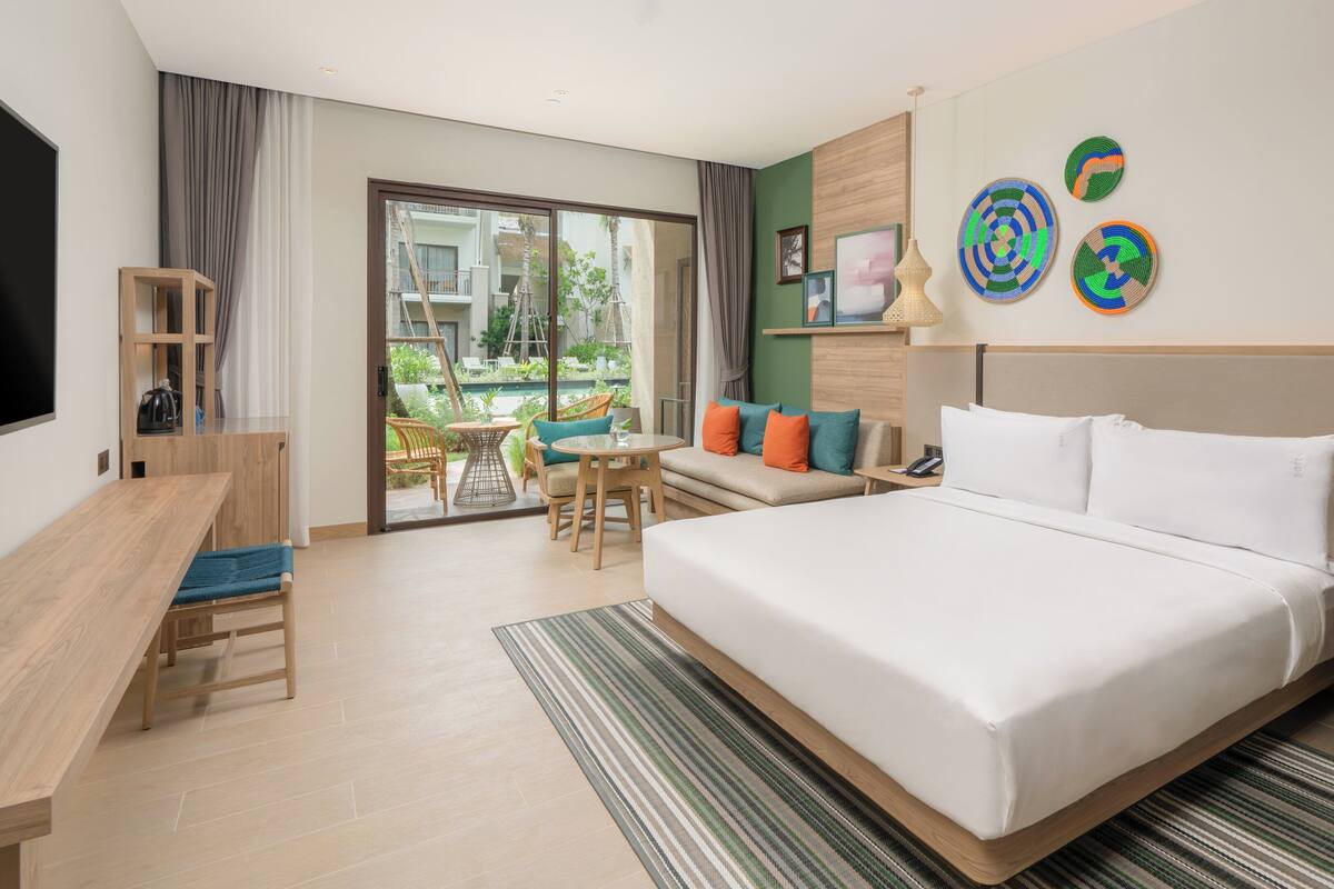 A hotel room with a large bed, a desk, a chair, a small table, and a view of the outdoor area.