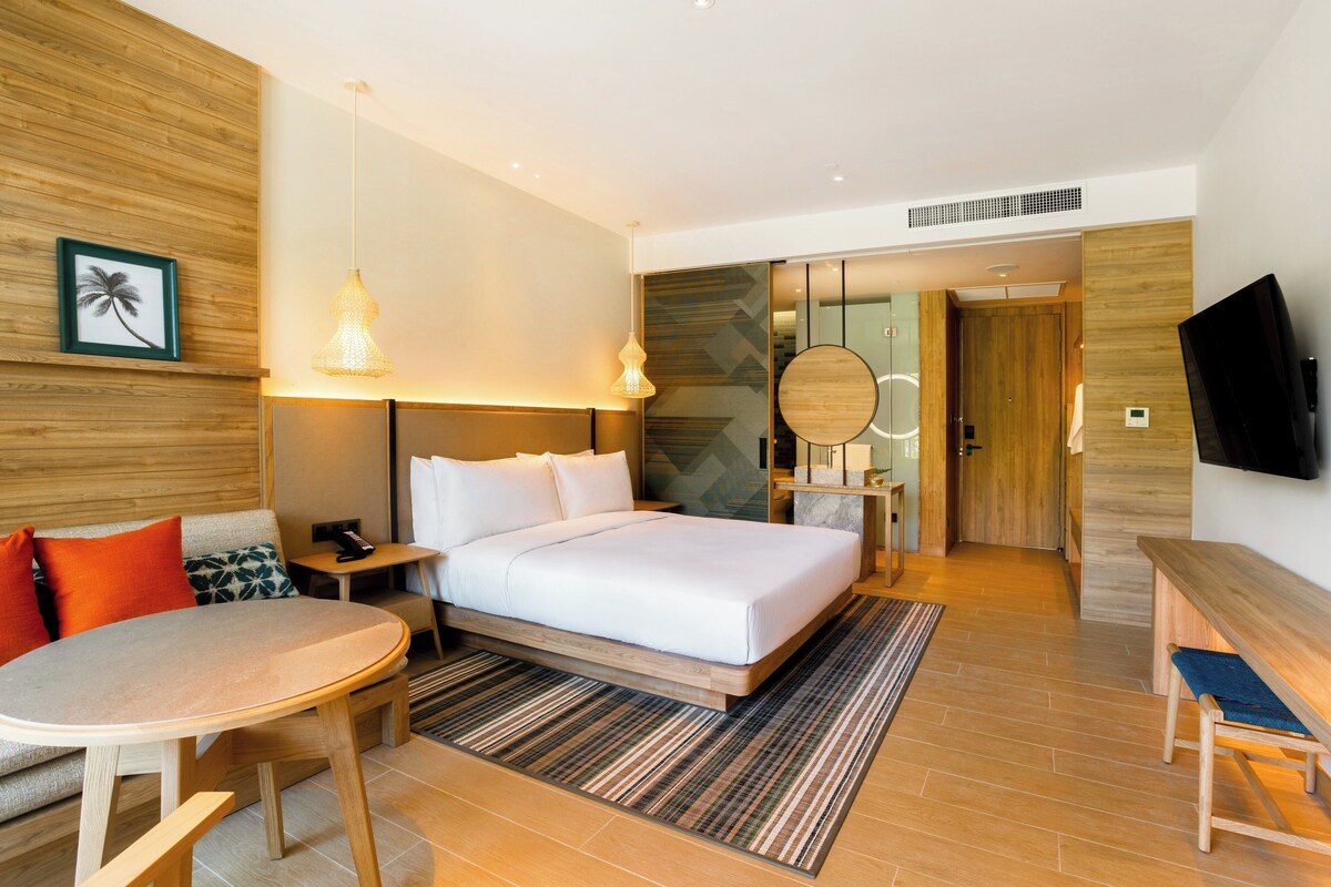 Una camera d'albergo moderna con un letto grande, uno specchio rotondo, una TV, una panca in legno, un tavolino e un quadro raffigurante una palma.