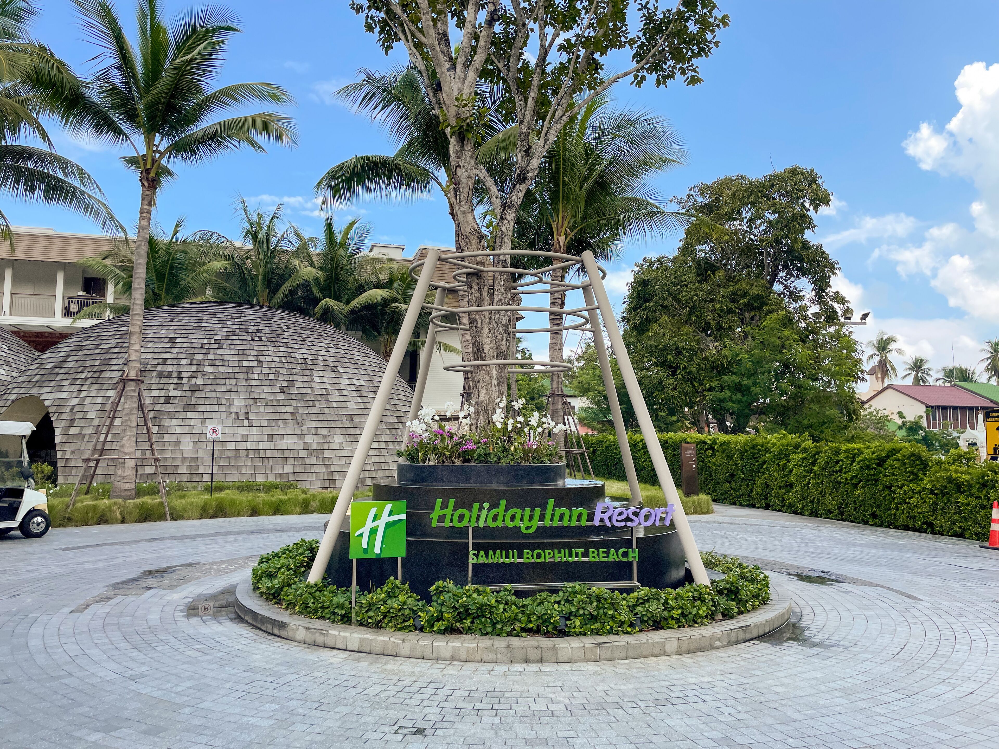 Une enseigne indiquant l’emplacement du Holiday Inn Resort, situé sur la plage de Samui Bophut, entouré de palmiers et bordé d’une zone pavée.