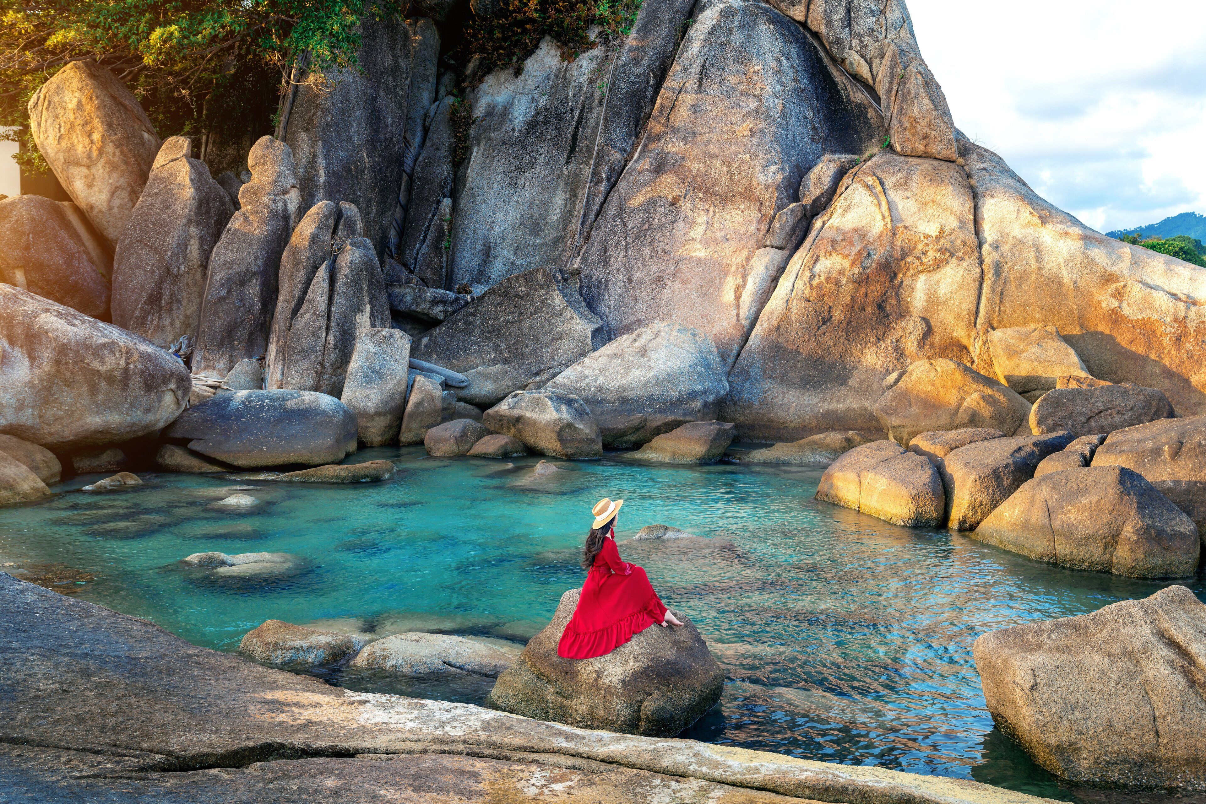 A woman in a red dress sitting on a rock by a clear water body surrounded by large rocks and a mountainous backdrop.