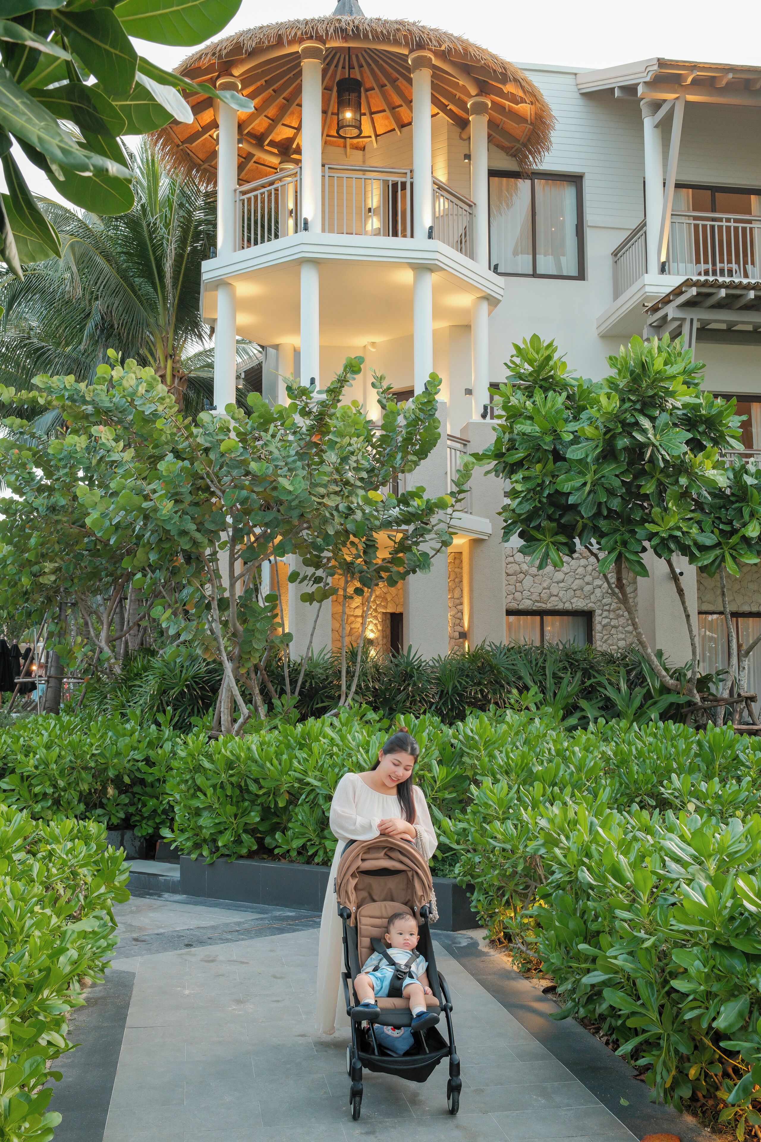 A woman pushing a stroller with a baby in a lush, green outdoor area with a building featuring a thatched roof and multiple balconies.