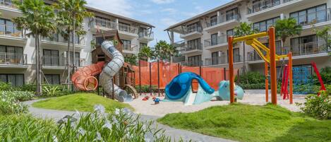 A playground with slides and climbing structures, surrounded by a landscaped garden and a pathway.