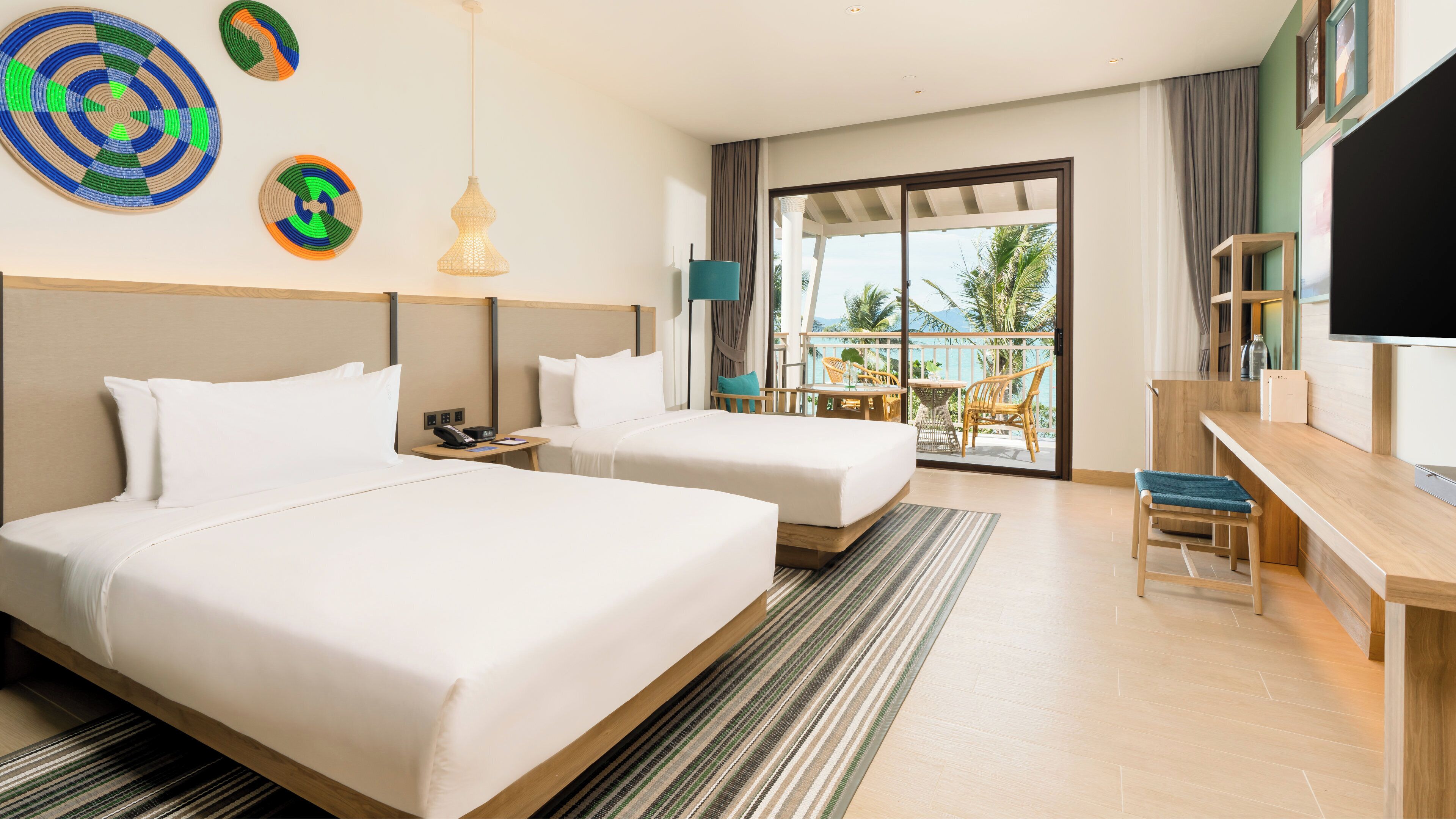 A hotel room with two beds, a desk, a TV, and a balcony with a view.