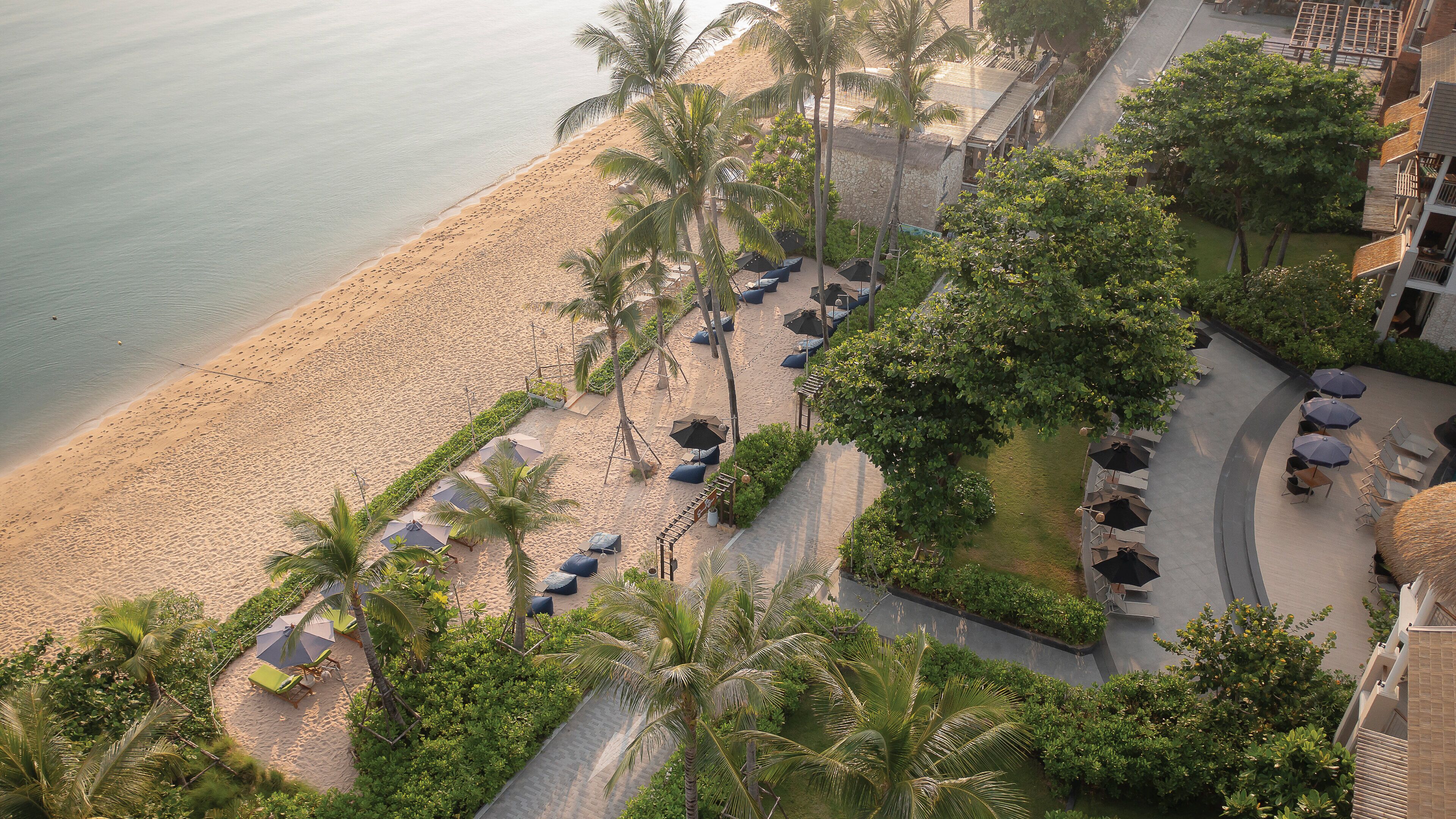 An aerial view of a resort with a beachfront, palm trees, and outdoor seating areas.