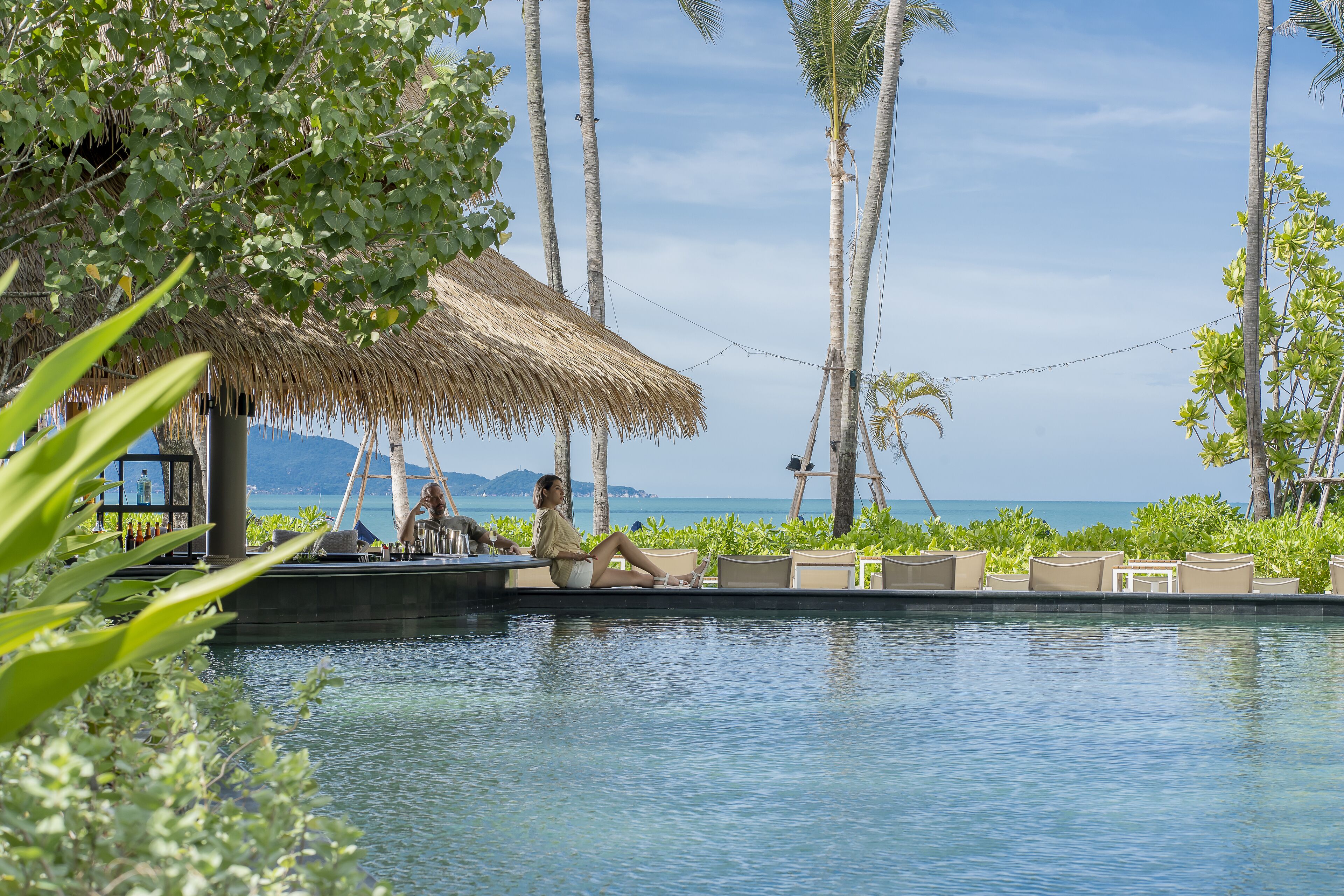 A poolside area with lounge chairs, a thatched-roof structure, and palm trees.