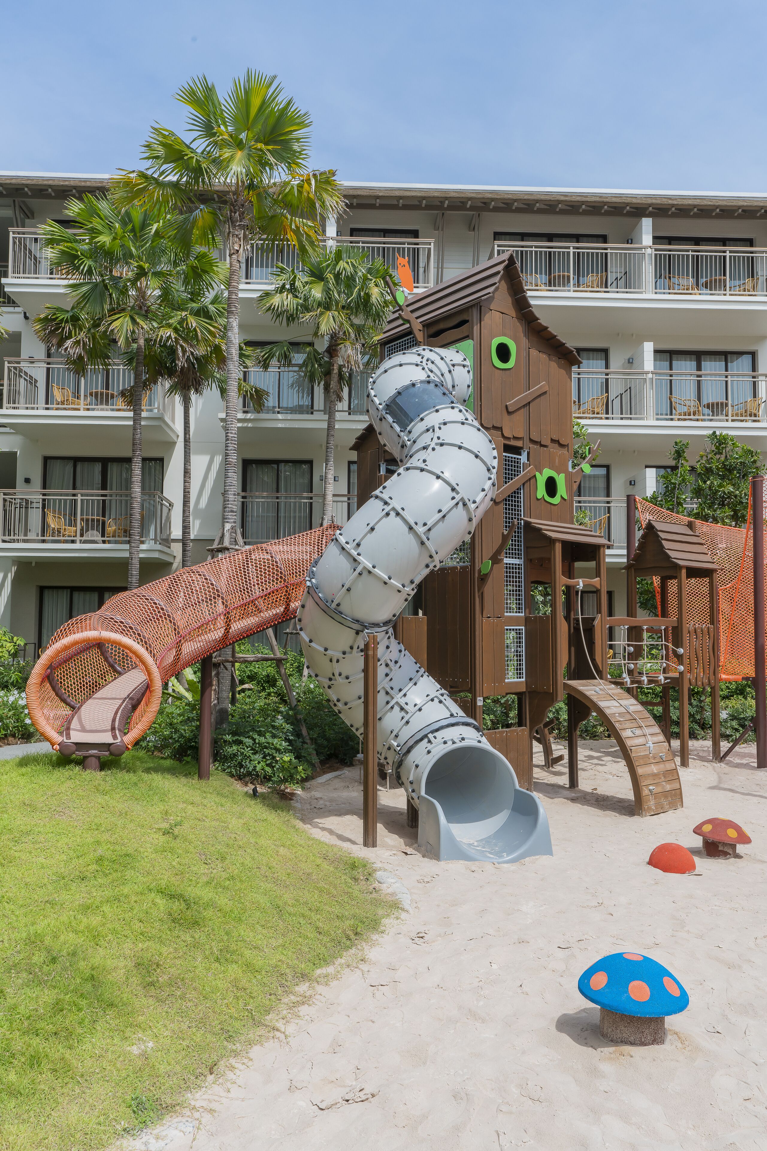 A playground with a slide and climbing structures, surrounded by palm trees and a building with balconies.