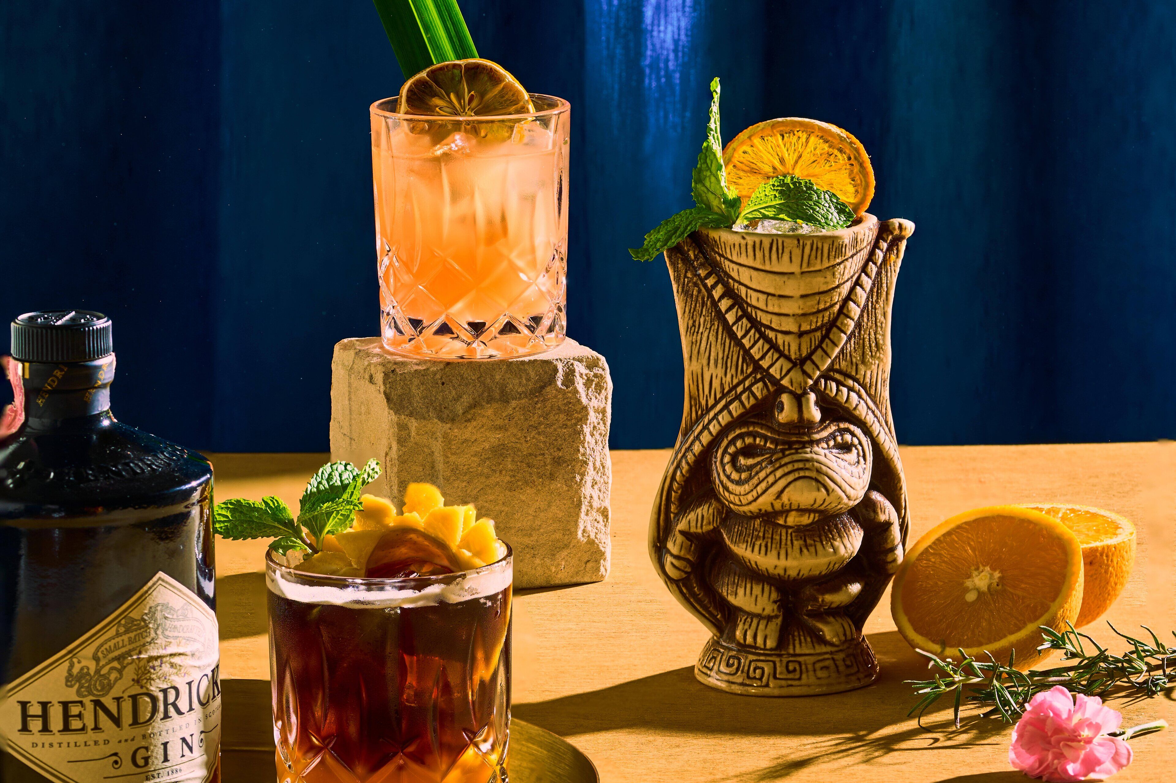 Eine Flasche Hendrick's Gin, ein Glas bernsteinfarbener Drink mit Zitronenscheibe, eine geschnitzte Holzvase mit einer halben Zitrone und eine halbe Orange auf einer Holztischplatte.