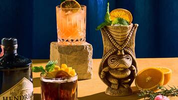 Eine Flasche Hendrick's Gin, ein Glas bernsteinfarbener Drink mit Zitronenscheibe, eine geschnitzte Holzvase mit einer halben Zitrone und eine halbe Orange auf einer Holztischplatte.