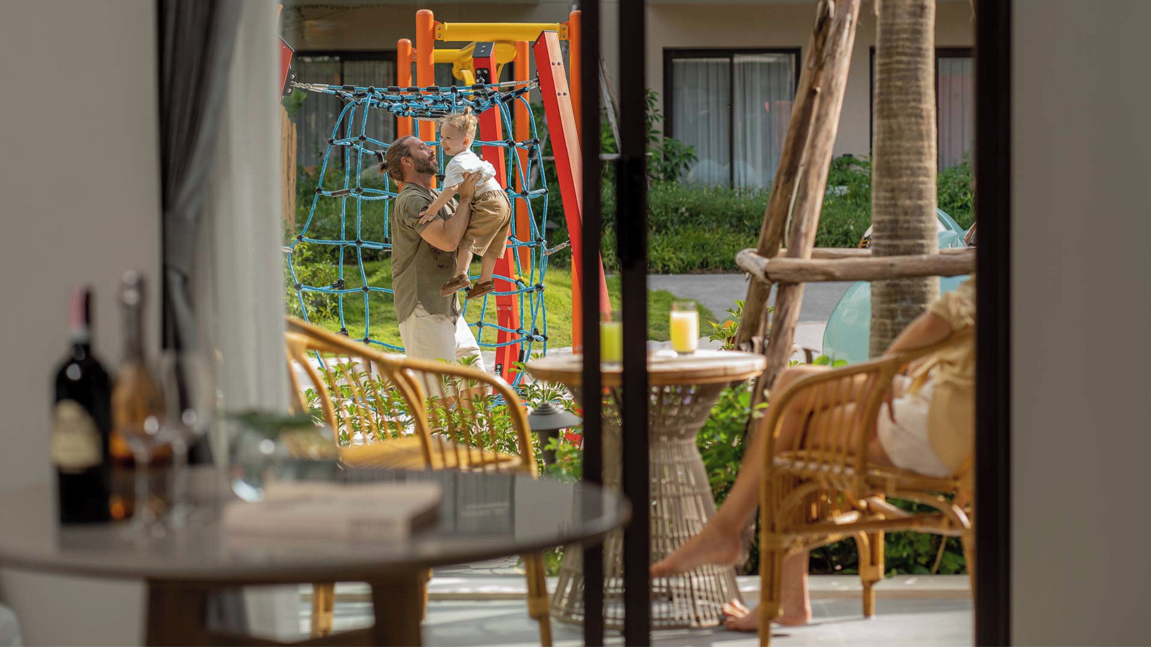 A man and a child on a rope bridge in an outdoor play area with a table and chairs nearby.