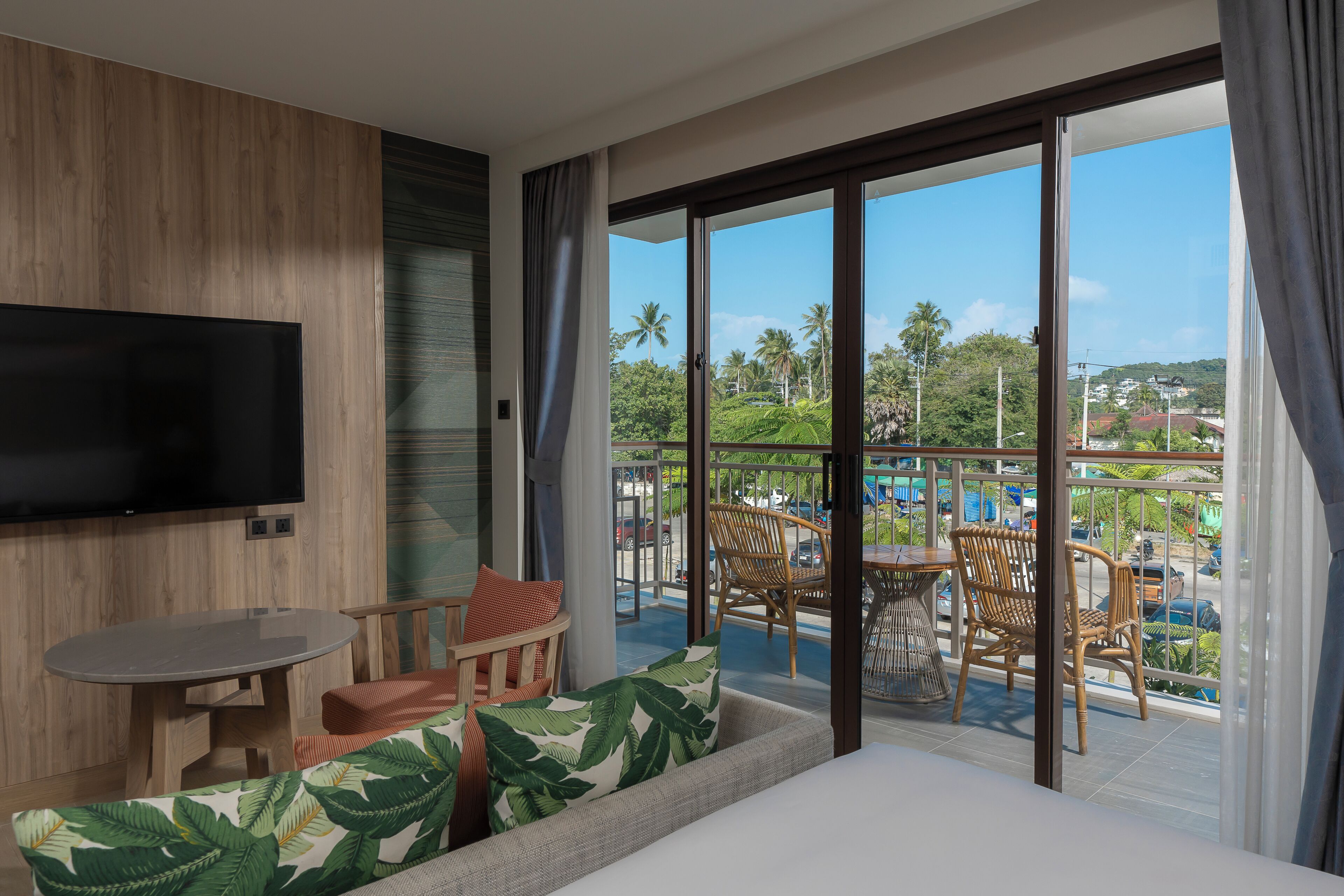 Habitación de hotel con balcón, televisión de pantalla plana, sofá con cojín estampado con motivos vegetales y mesa de comedor con sillas.