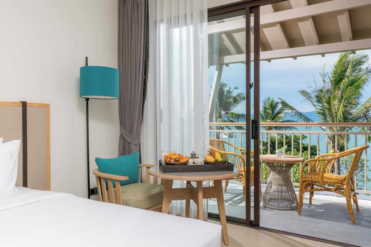 Camera d'hotel con un letto, una sedia, un tavolo con frutta e un balcone vista mare.