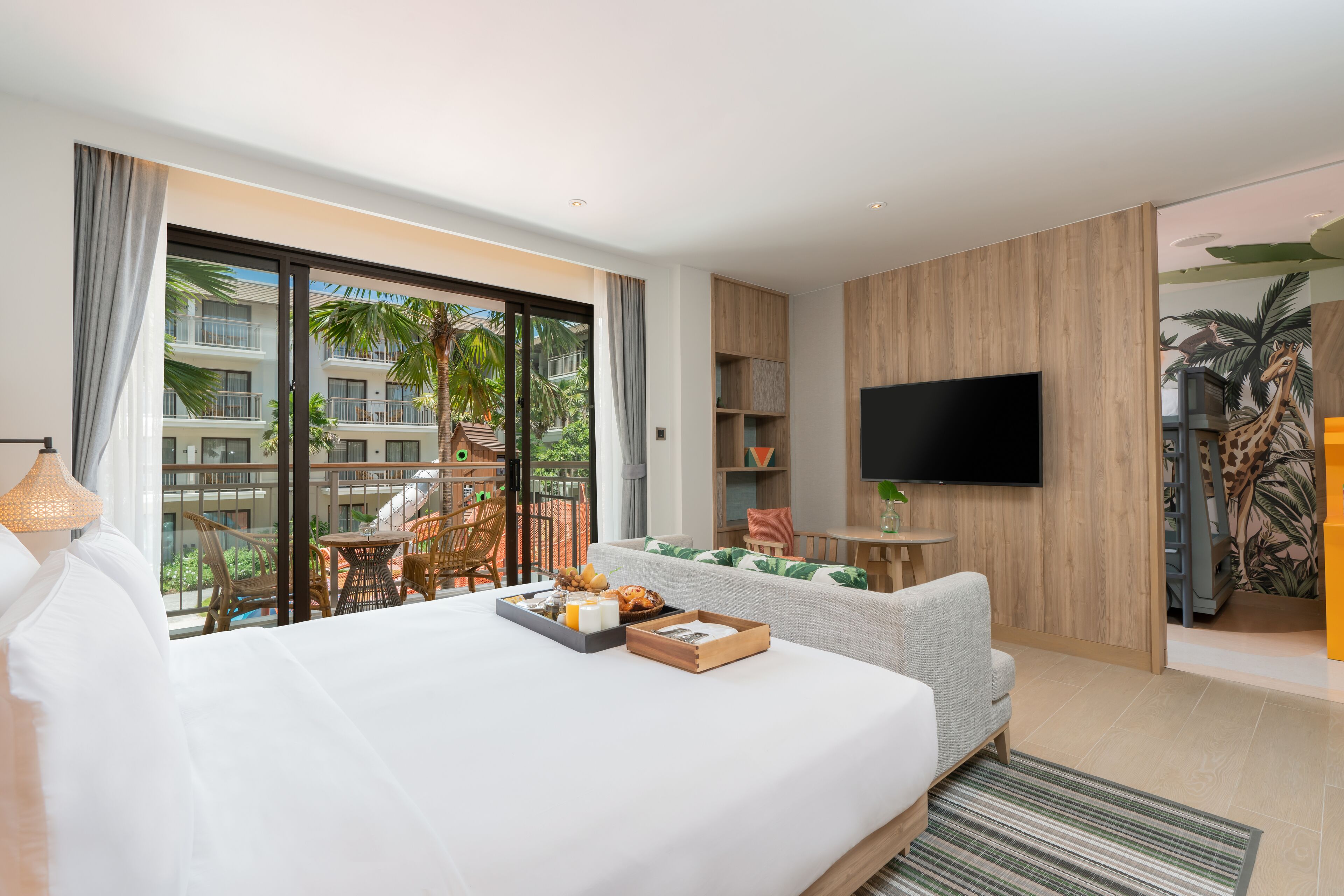 Een moderne hotelkamer met een groot bed, een televisie, een balkon met tuinmeubilair en uitzicht op een gebouw met palmbomen.