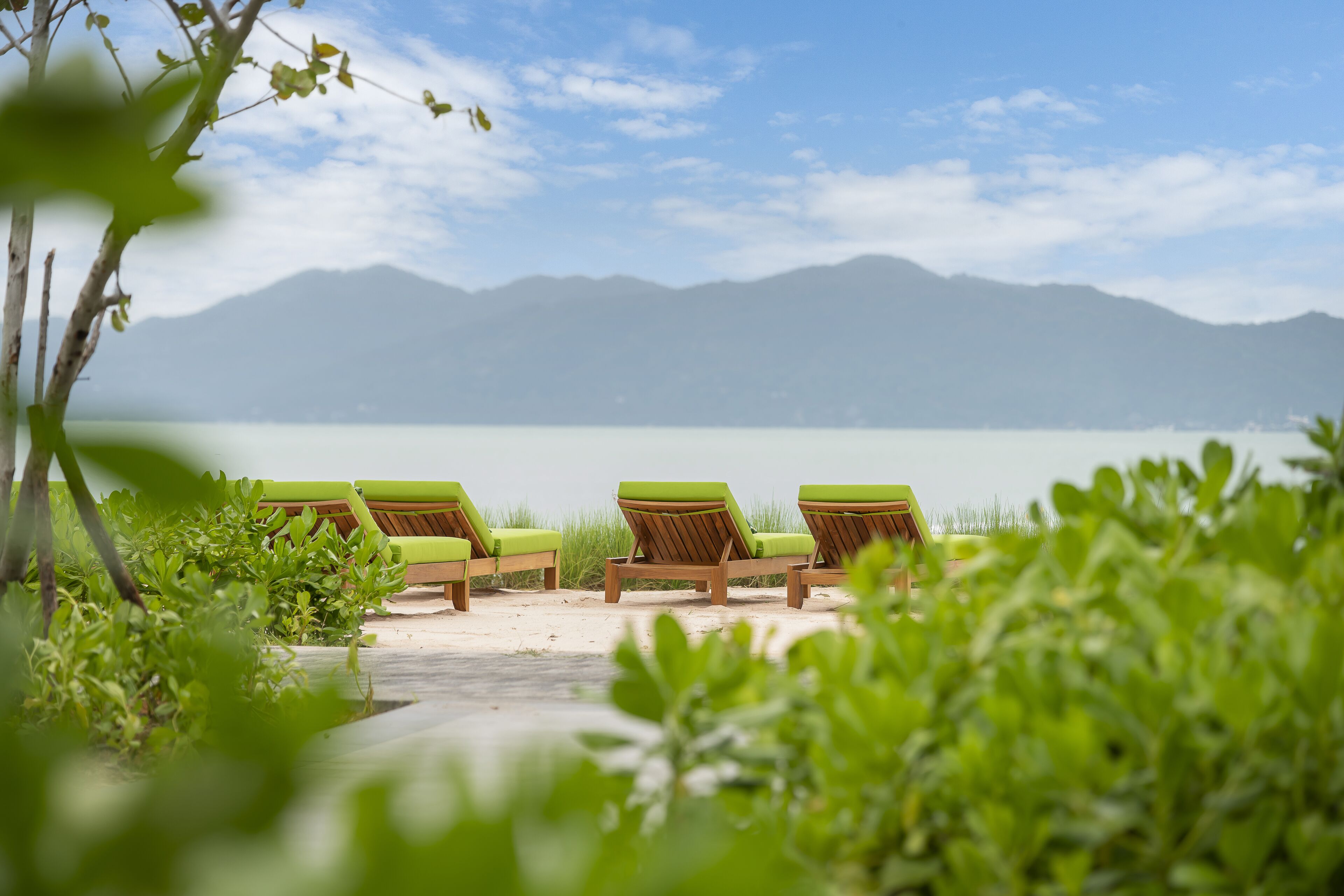 Una hilera de sillones de madera con cojines verdes sobre una playa arenosa, con vistas a un lago tranquilo y montañas a lo lejos.