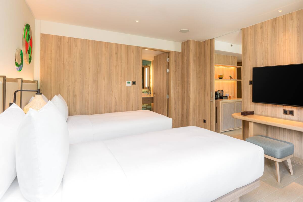 A modern hotel room with two beds, a TV mounted on the wall, and a kitchenette in the background.