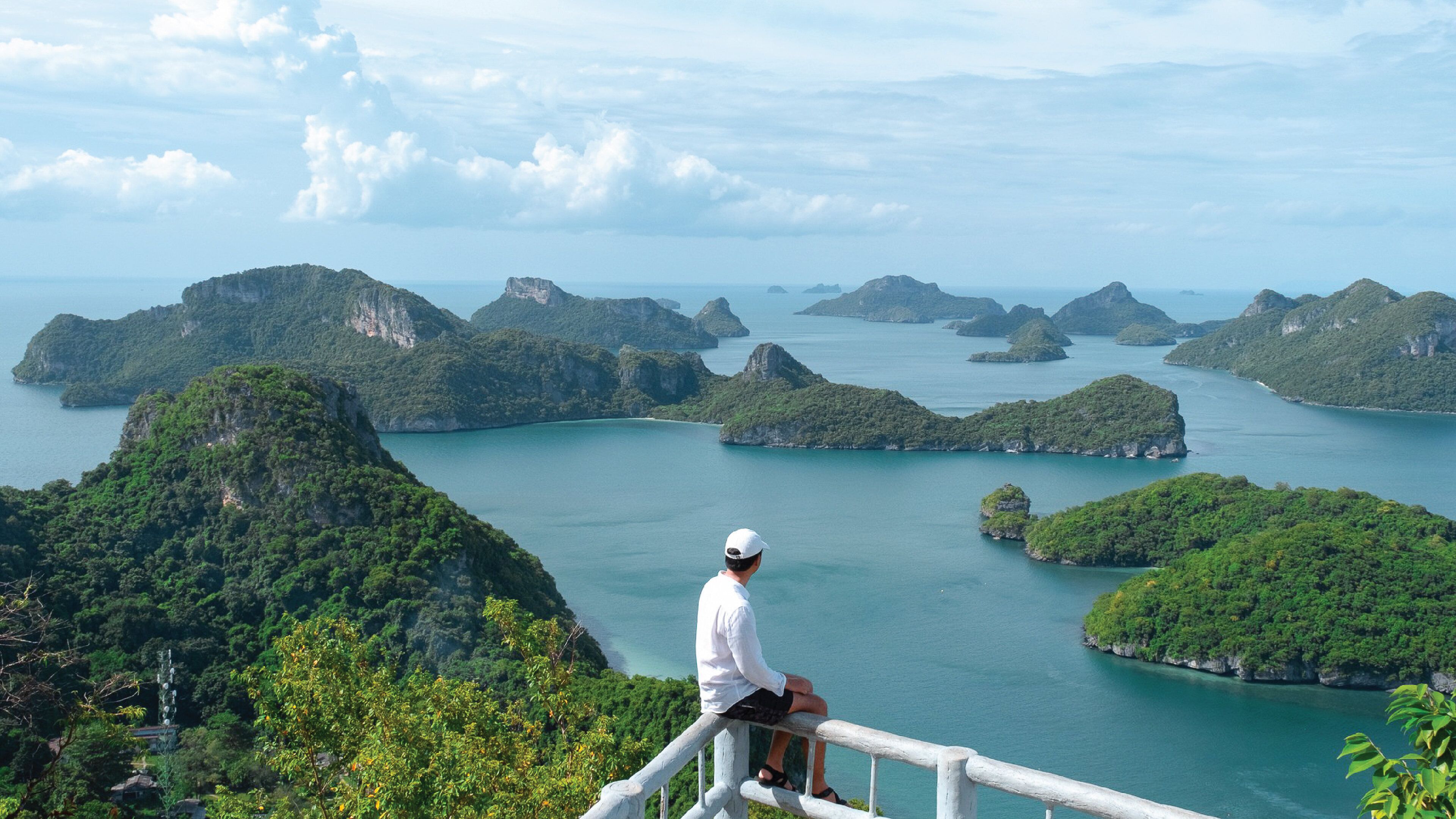 A person sitting on a railing overlooking a coastal landscape with numerous islands and a clear sky.
