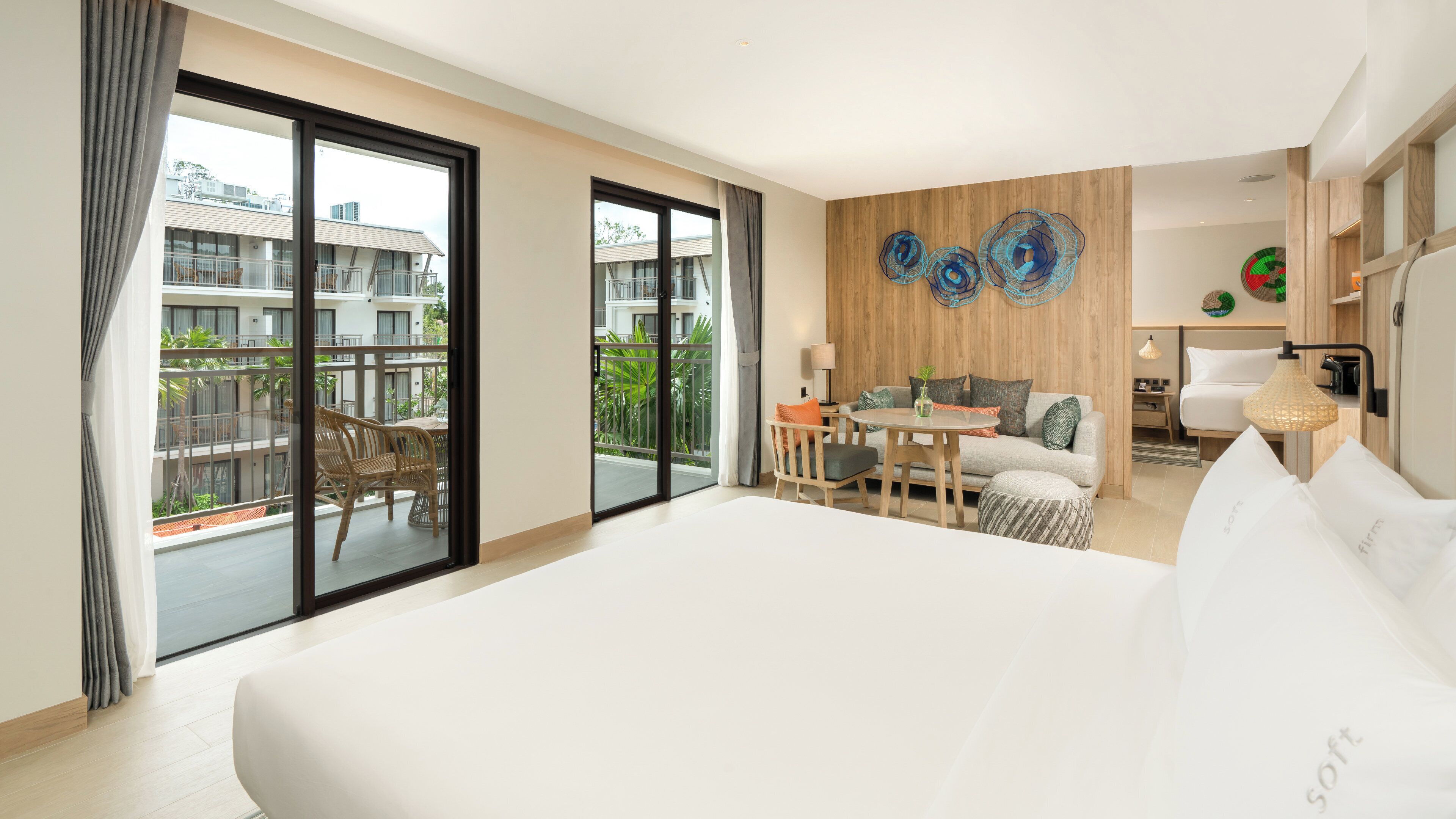 A hotel room with a large bed, a balcony with wicker furniture, a wooden accent wall, and a dining area with a table and chairs.