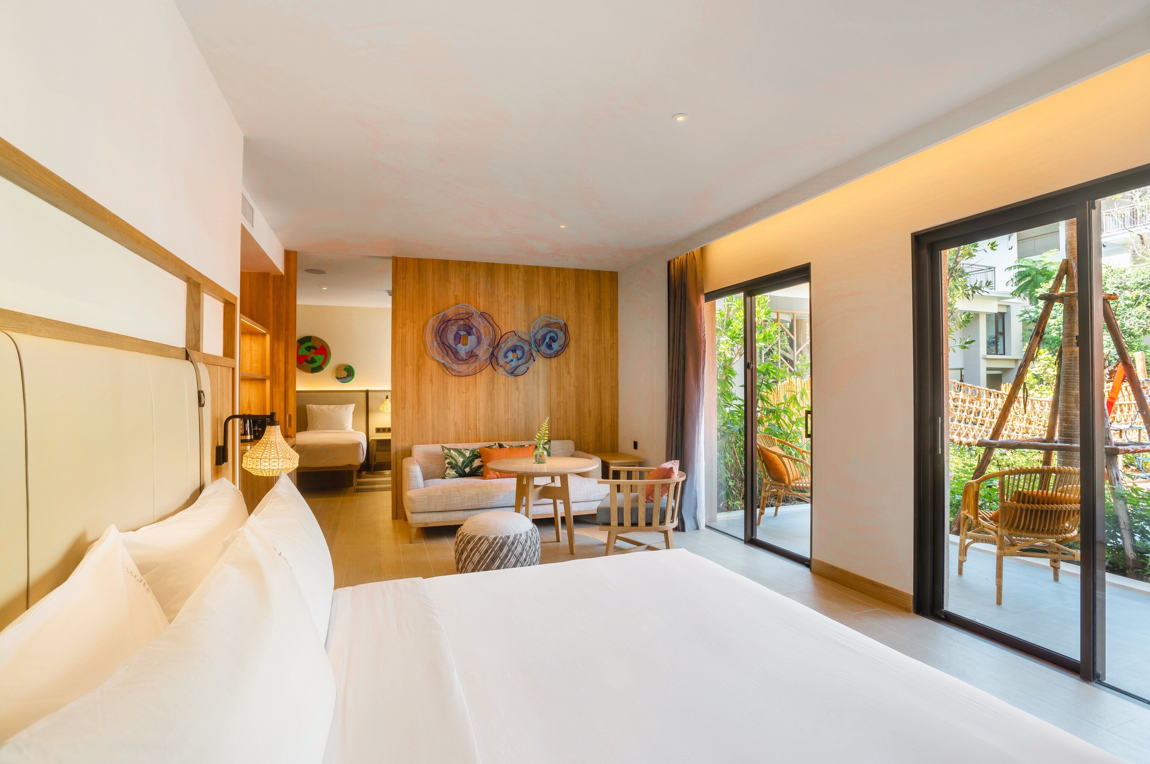 A modern hotel room with a large bed, a sofa, a dining table, and a balcony with wicker furniture.