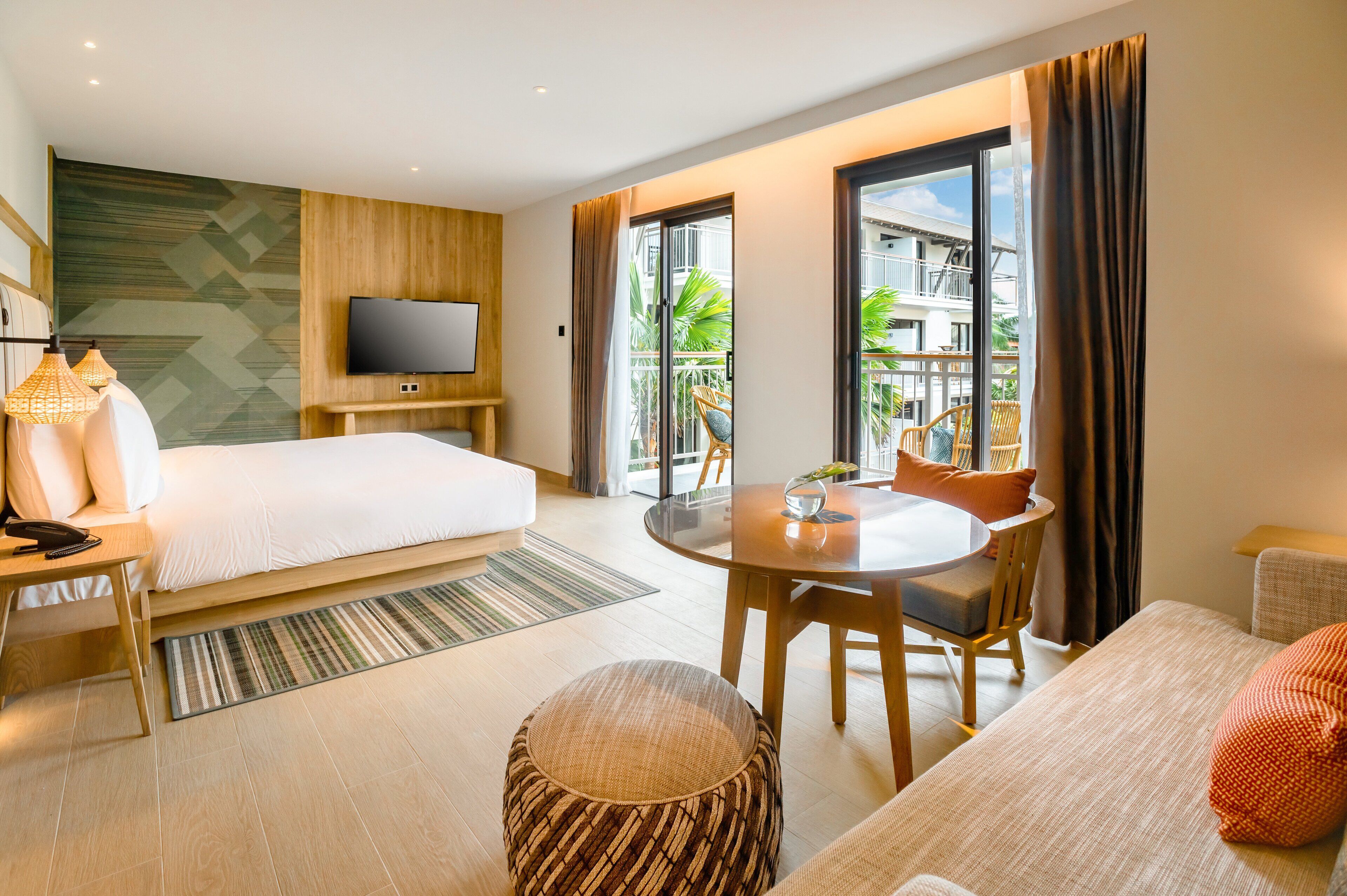 A hotel room with a bed, a TV, a dining table with chairs, and a balcony with a view of buildings and plants.