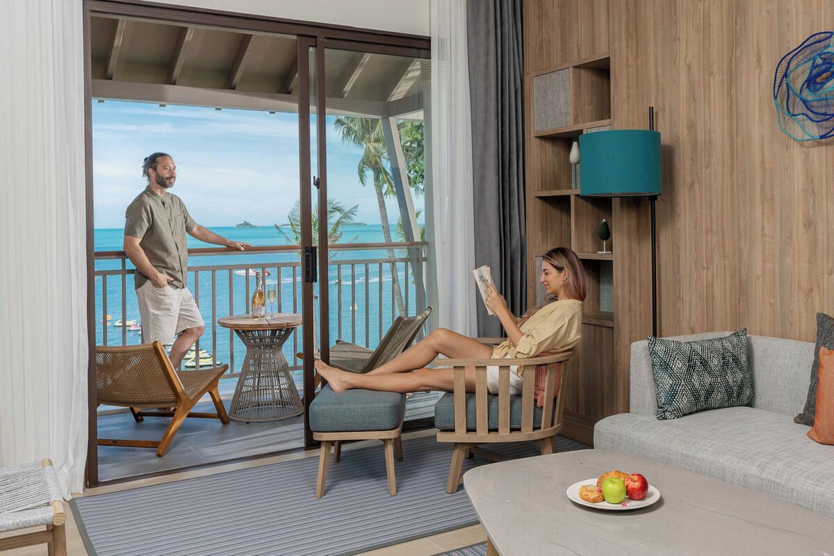 Una stanza con un balcone vista spiaggia, un divano con cuscini, un tavolino con un piatto di frutta e una libreria.