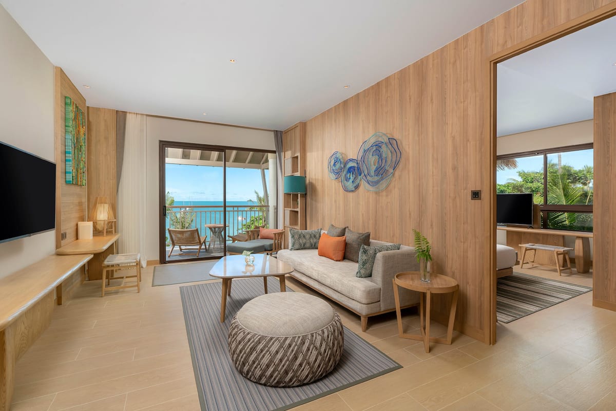 A modern living room with a sofa, coffee table, and ottoman, a flat-screen TV, and a large window offering a view of the ocean.