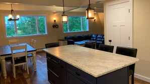 A kitchen with a central island, dark cabinetry, and pendant lighting. There are windows with views of greenery, a white door, and a black leather bench in the background.