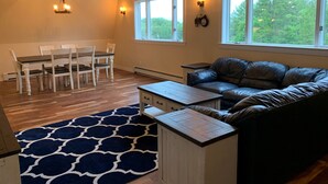 A spacious living room with a dining area, a leather sofa, a patterned rug, and large windows offering a view of trees.