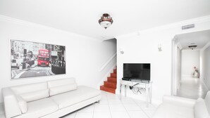 A modern living room with a white sofa, a flat-screen TV on a white stand, a staircase with red steps, and a large black-and-white cityscape painting on the wall. - Beautiful 2/2.5 Condo w/Free Parking Fast WIFI (Miami)