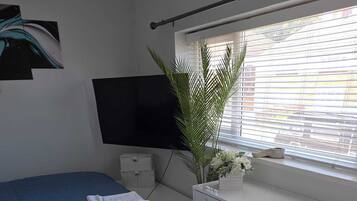 A bedroom with a bed, a desk, a television, a window with blinds, and a potted plant.