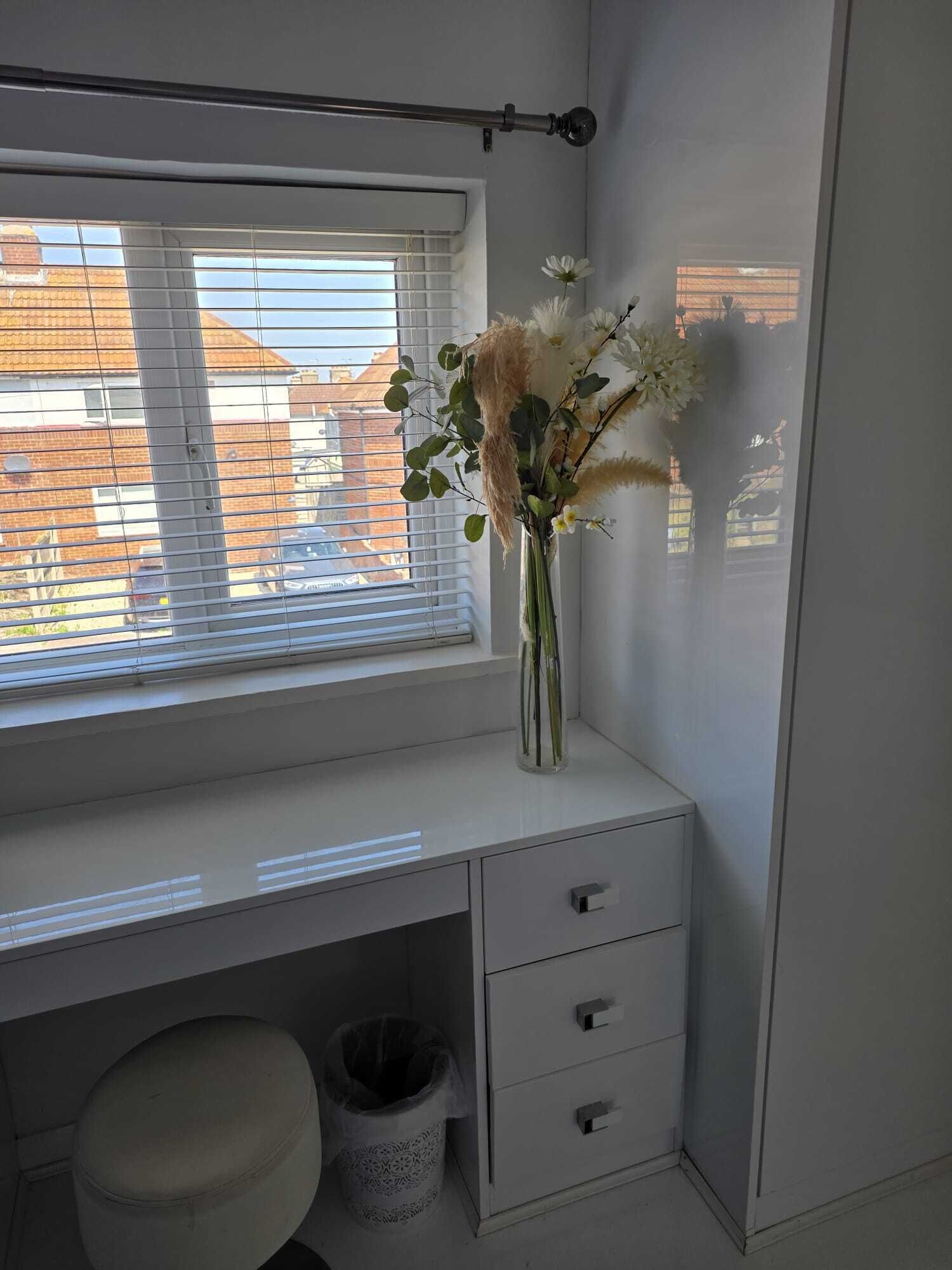 Ein Zimmer mit Fenster, Schreibtisch mit Schubladen, Mülleimer und ein kleiner Hocker.