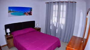 A bedroom with a bed, nightstands, a wooden dresser, a painting of a beach scene, and a window with curtains. - Suitable. Deep beach, a few meters from the beach. (Playa Honda)