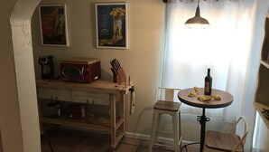 Une cuisine chaleureuse avec une table en bois, des chaises, un four à micro-ondes et une machine à café. Deux tableaux encadrés ornent le mur, et une fenêtre est agrémentée de rideaux.