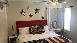 Une chambre avec une tête de lit rouge, du linge de lit blanc et une couverture ornée du drapeau américain. Des décorations en forme d’étoiles ornent le mur et un ventilateur de plafond est installé au-dessus du lit.