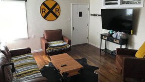 Un salon avec un canapé en cuir marron, une table basse en bois, un ventilateur de plafond, un téléviseur et un panneau décoratif fixé au mur.