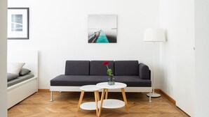 Un salon moderne avec un canapé gris foncé, deux petites tables rondes blanches, un parquet en bois disposé en chevrons, un tableau encadré au mur et un luminaire suspendu.
