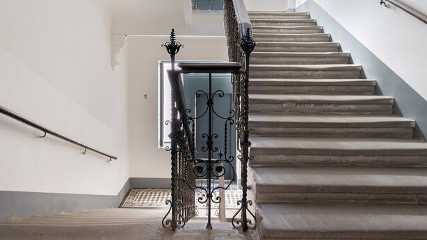 Un escalier dont le sol présente un motif à carreaux, avec une rampe en métal, et qui offre une vue sur une porte située en haut.