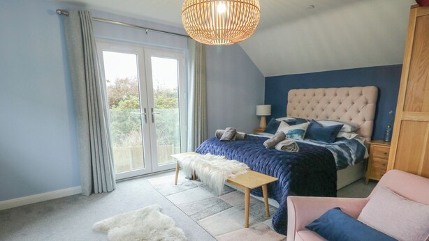 A bedroom with a large bed, a pink chair, a white furry rug, and a wooden nightstand. - 11 Parc Delfryn (Brynteg)