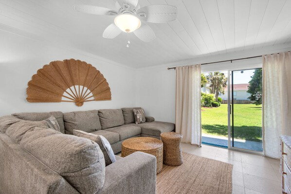 Ein Wohnzimmer mit einem Ecksofa, einem Deckenventilator, einem gewebten Beistelltisch und Blick durch eine Schiebetür.