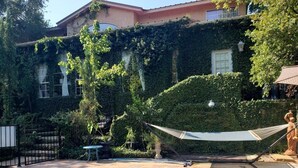 A swimming pool with a hammock and a statue, surrounded by greenery and a building with ivy-covered walls.