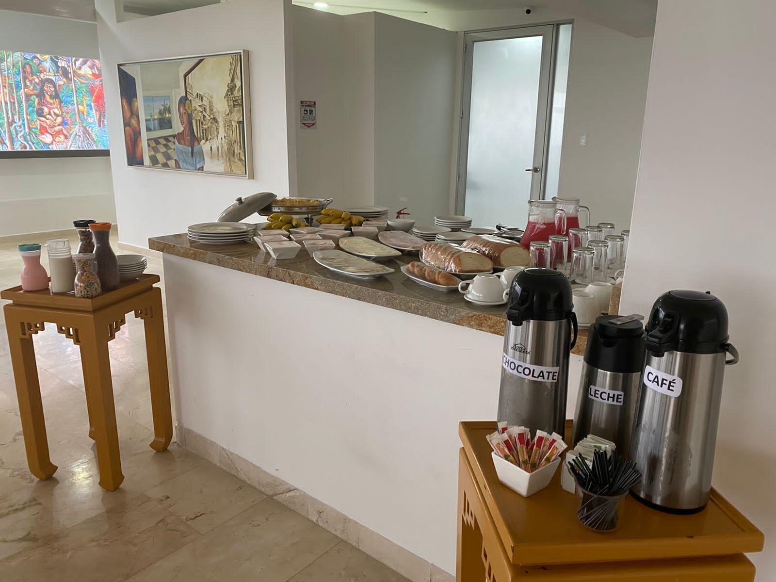 Un buffet de petit-déjeuner proposant divers aliments et boissons, un tableau accroché au mur et une petite table avec des condiments.