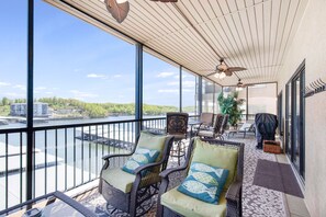 A balcony with wicker chairs, a table, and a view of a river and buildings. - Amazing Main Channel Views with Screened Balcony (Lake Ozark)