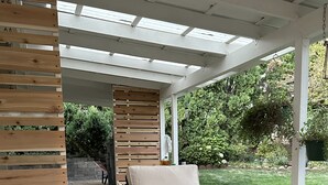 En udendørs patio med fletmøbler, et træbord og en pergola med hvidt tag.