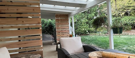 A covered patio area with wicker furniture and a wooden table, overlooking a garden.