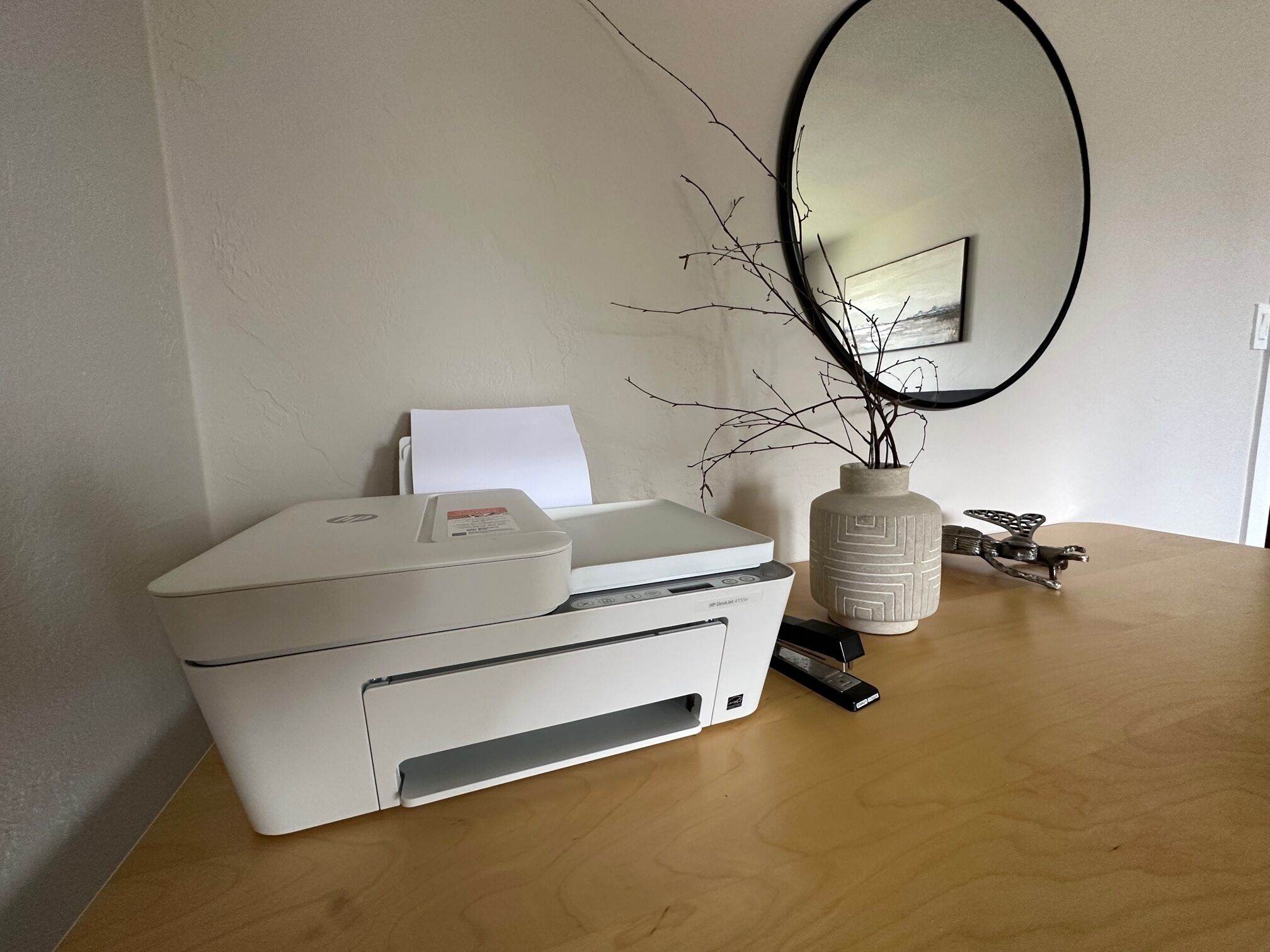 Une imprimante posée sur une table en bois, avec un vase rempli de branches et un miroir accroché au mur.