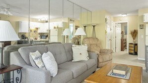 Un salon avec un canapé gris, une table basse en bois et un fauteuil beige. On peut apercevoir une cuisine à travers une cloison en verre.