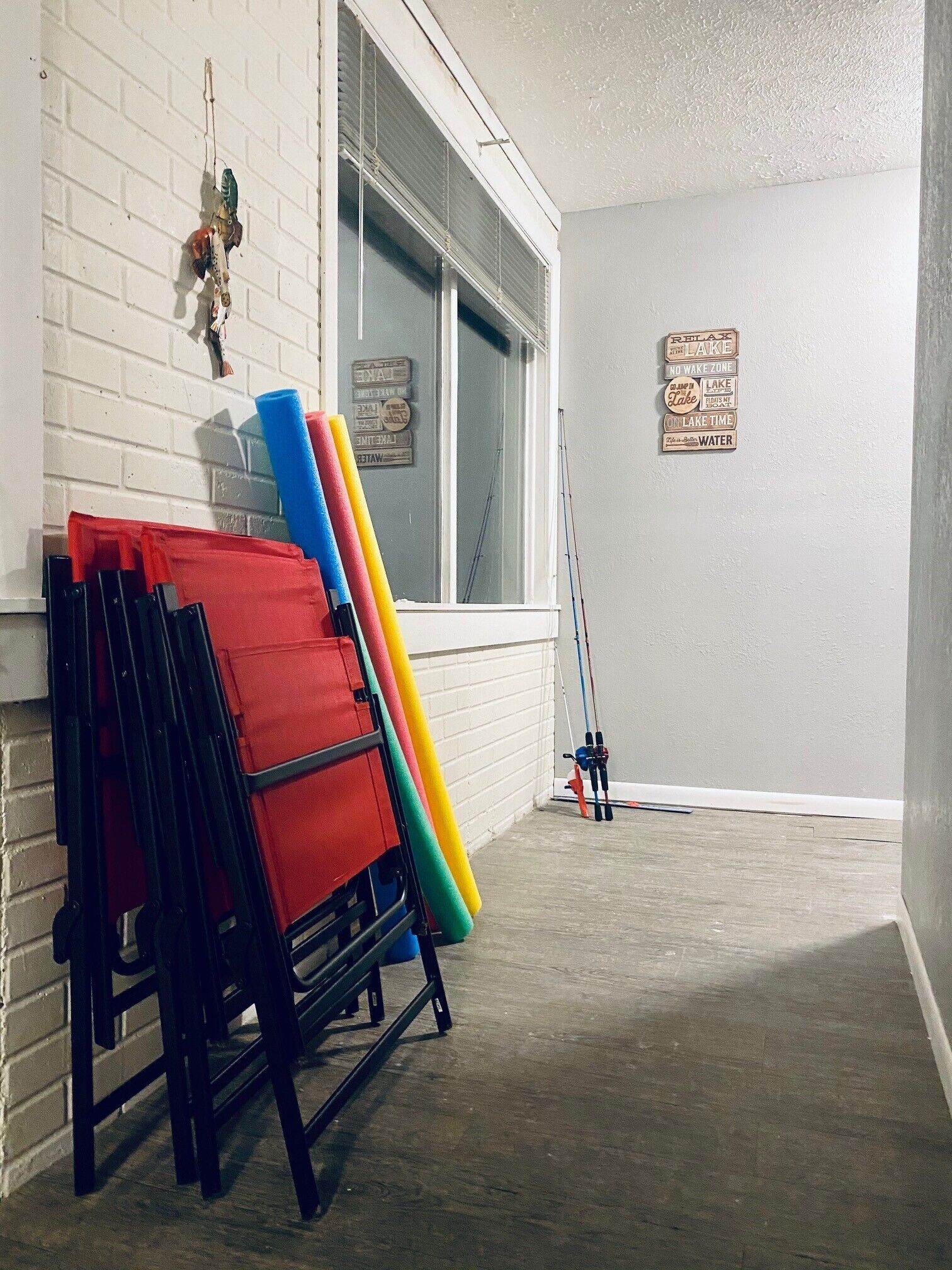Une pièce avec un mur en briques blanches, une fenêtre avec des stores, un panneau fixé au mur et une chaise pliante agrémentée de tapis colorés.