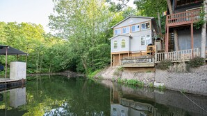Une maison à deux étages dotée d’un balcon donnant sur un lac paisible, le tout entouré d’une végétation luxuriante.