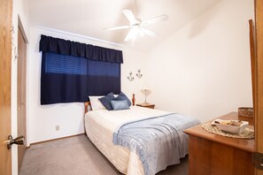 Une chambre à coucher comprenant un lit, une table de chevet, un bureau, un ventilateur de plafond et une fenêtre avec des stores.