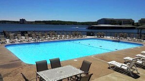 A rectangular pool with lounge chairs and tables, surrounded by a fence, with a view of a body of water and buildings in the background. - Amazing Lakefront Pool On Coveted Horseshoe Bend! (Four Seasons)