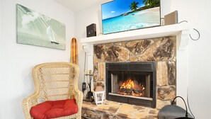 Een woonkamer met een stenen openhaard, een rieten fauteuil met een rode kussens en een flatscreen-tv op een schoorsteenmantel, en een schilderij van een zeilboot.