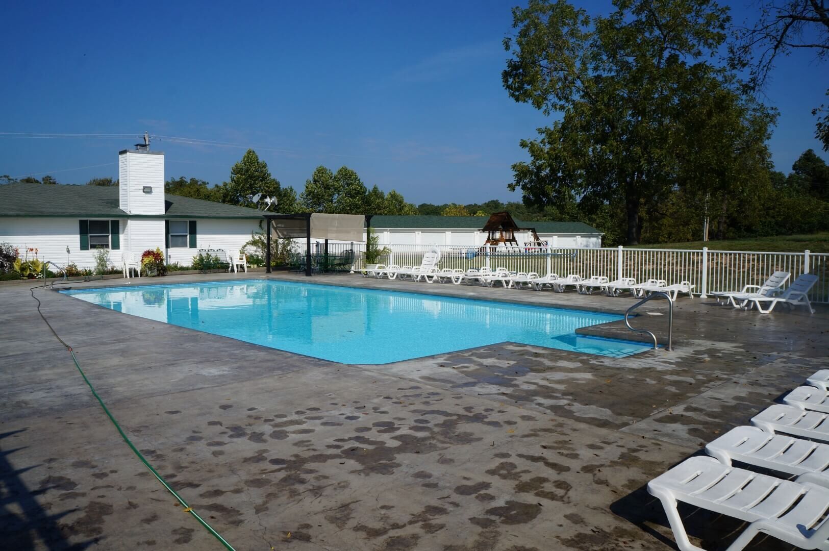 A swimming pool with a concrete deck, surrounded by white lounge chairs and a covered area with a roof.